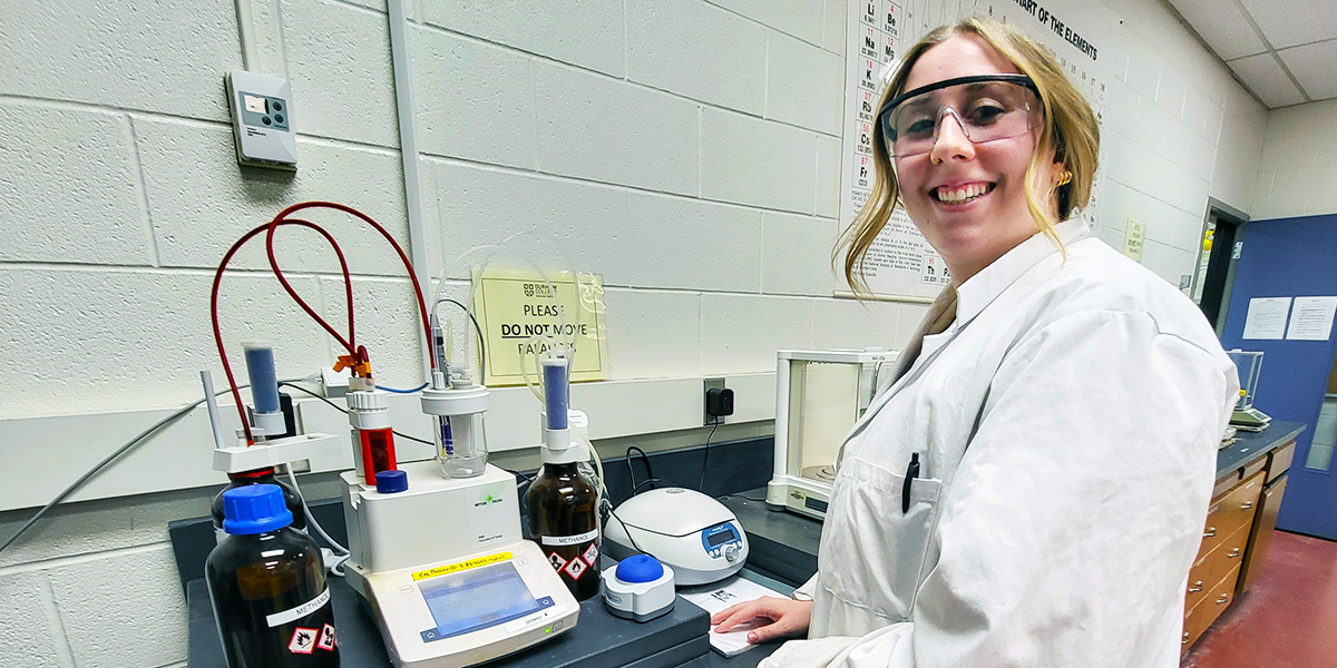 In a white lab coat, Melanie Williams stands in front of scientific equipment and smiles.