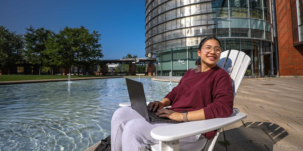 A students sits outside the Durham College Library with a laptop open in her lap.