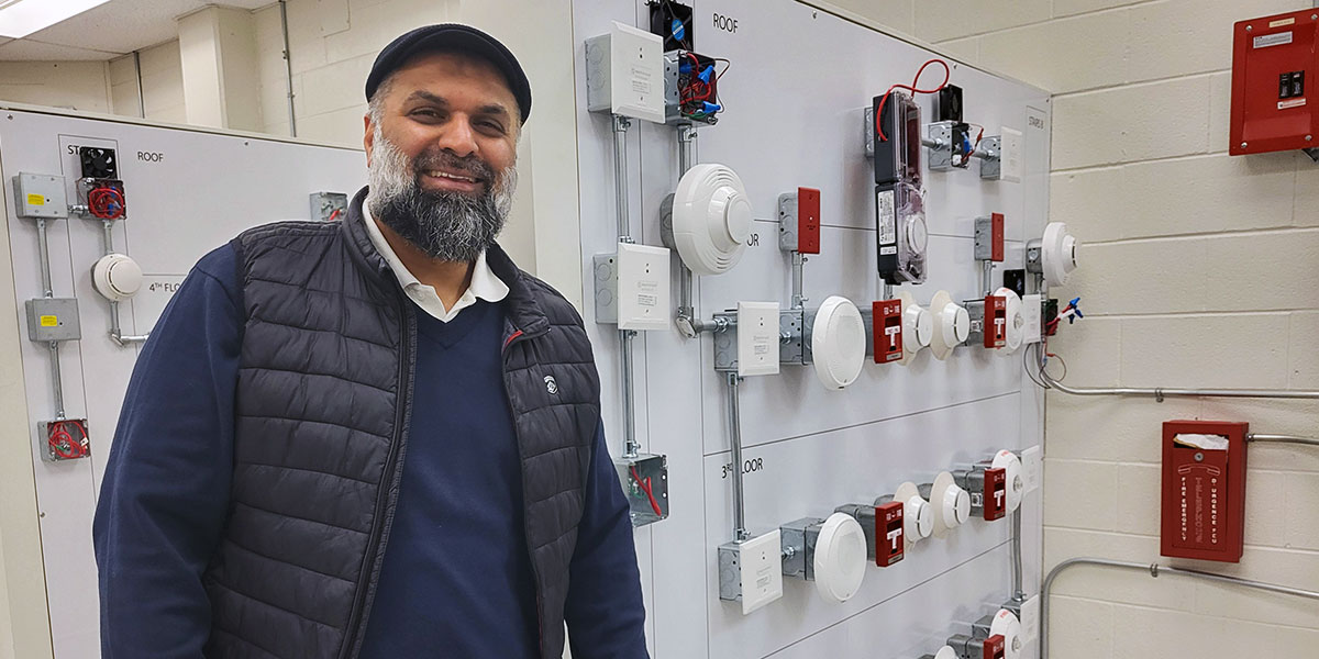Randy stands beside a wall-mounted display of fire alarm system components,