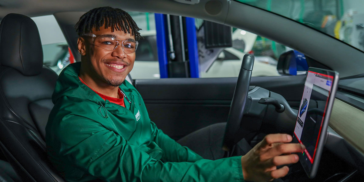 A smiling person wearing glasses and a green jacket sits in the driver’s seat of a car, using the vehicle’s touchscreen display, with a garage or service area visible in the background