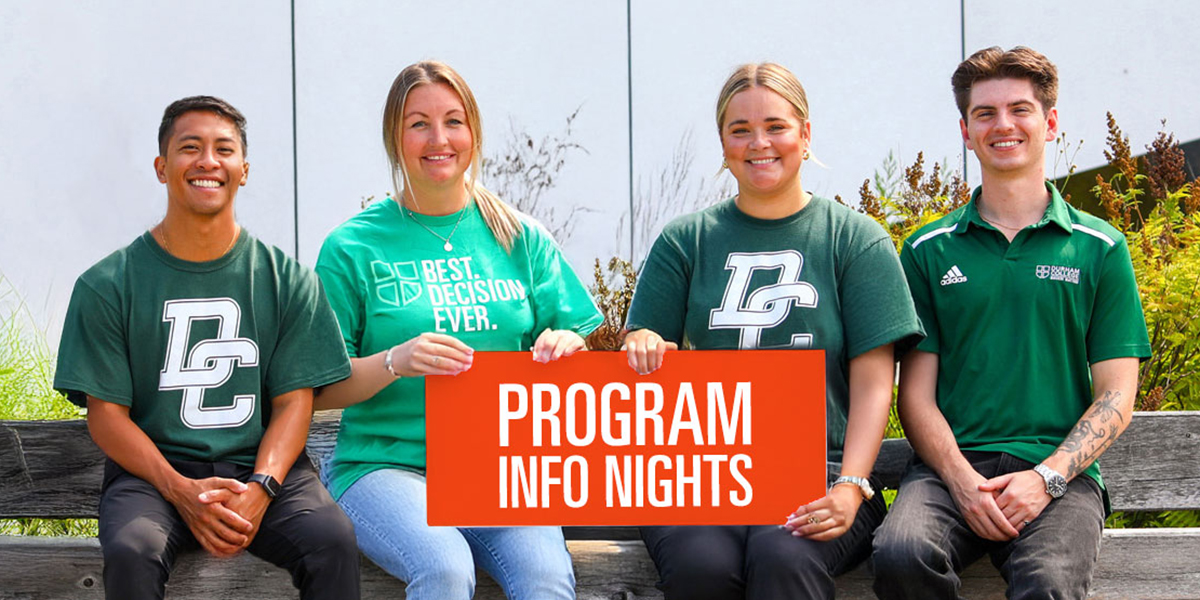 Four students sit side by side outdoors on a bench, smiling and holding a bright orange sign that reads “Program Info Nights.” They are wearing green college-branded shirts, suggesting school pride, with a modern building and plants in the background.