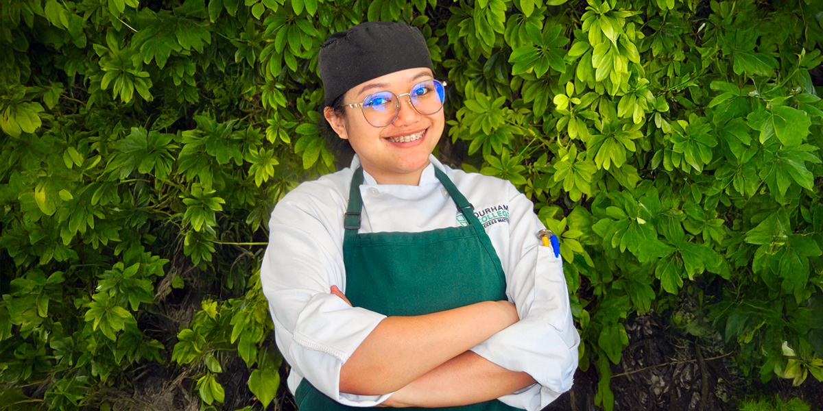A smiling culinary student in a chef’s uniform, apron, and glasses stands with arms crossed in front of a leafy green wall.