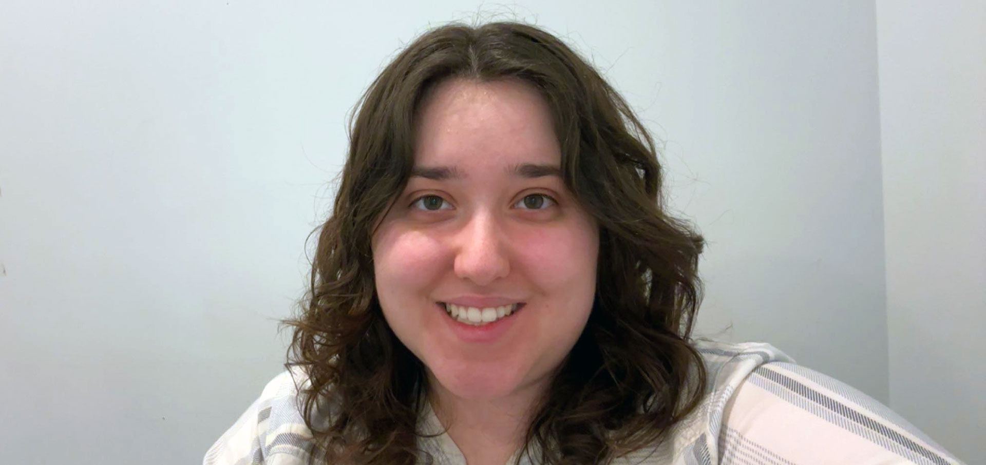 Adrianna smiles at the camera while seated indoors against a light-colored wall.