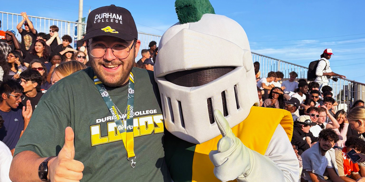 A man wearing a Durham College Lords shirt and cap smiles and gives a thumbs-up while posing beside a knight mascot in the stands at an outdoor sporting event.