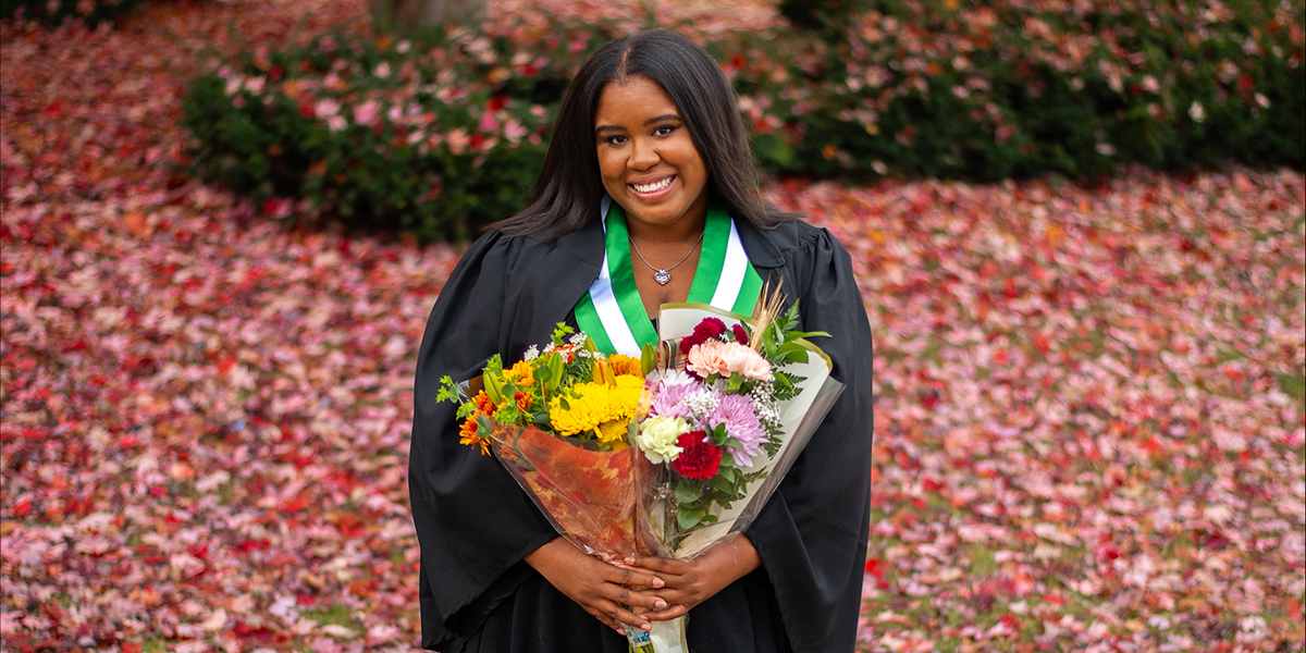 Reagan wears a black graduation gown and a green-and-white stole and stands outdoors holding two bouquets of flowers. Red and pink autumn leaves cover the ground and fill the background.