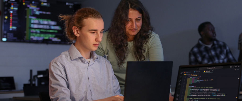 Faculty member assisting a student on a laptop in a computer lab.