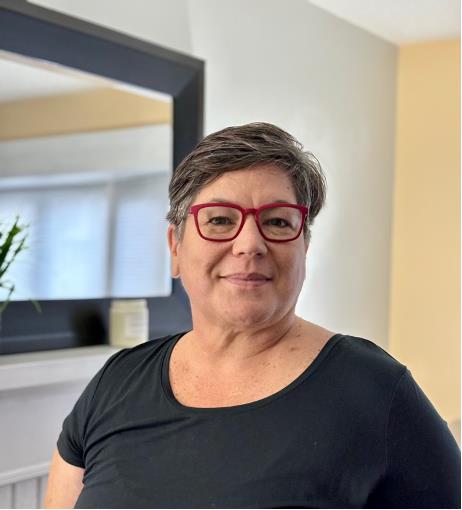 Ela Smith - a woman with short greying hair and red glasses wearing a black shirt in front of a white wall with a mirror.