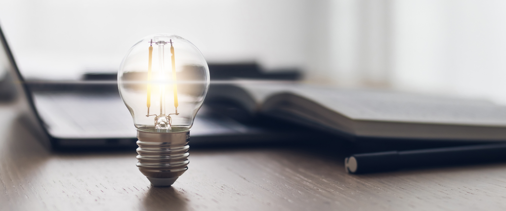 Lightbulb sitting on a desk with an open notebook and laptop.