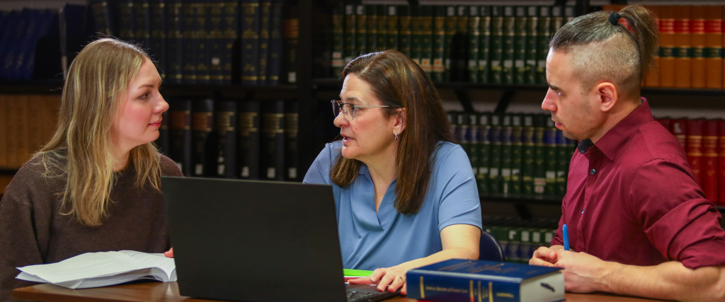 Faculty member speaking to two students in front of several bookshelves.