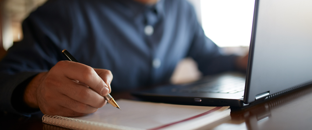 Student sitting in front of a laptop and writing on an open notebook.
