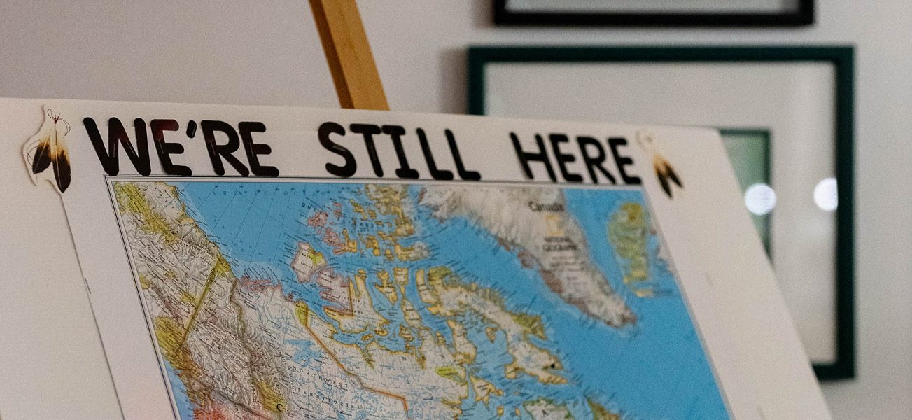 Map of Canada on an easel with "We're Still Here" across the top