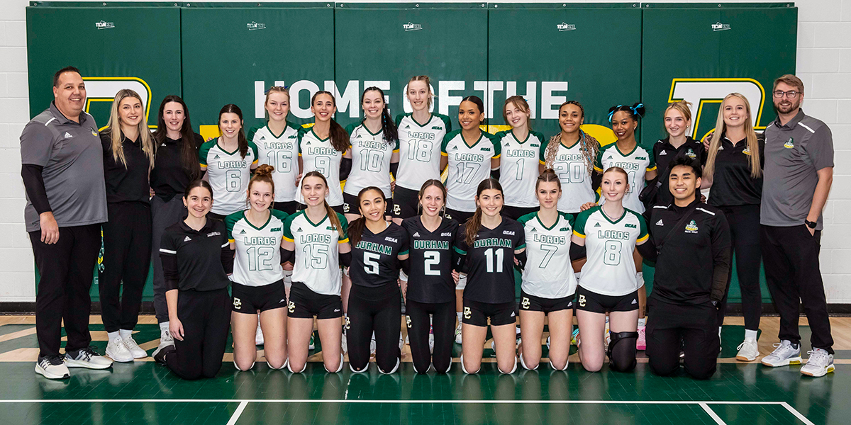 Durham College's women's volleyball team gathers for a group photo wearing their white, green, and black volleyball uniforms with their coaches standing on at each end of the group.