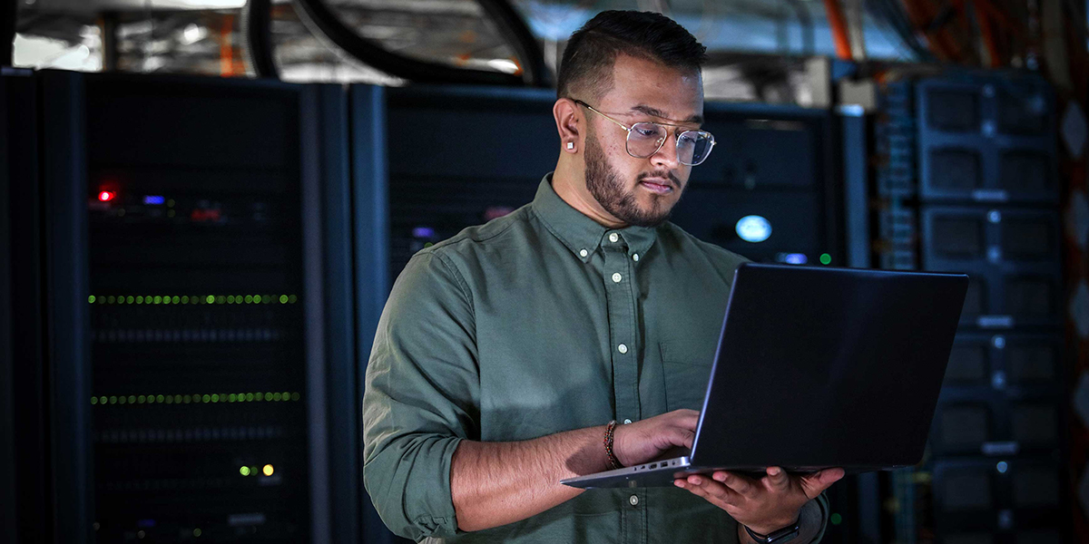 A man in a computer room consults his laptop.