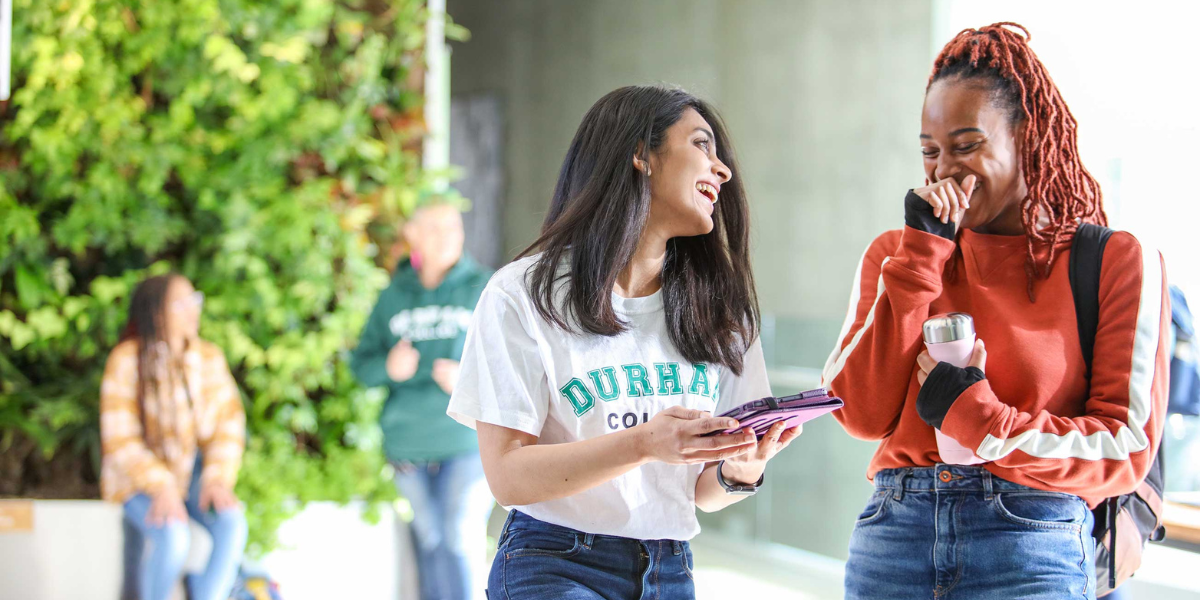 Two students stand inside a Durham College building laughing while looking at a tablet.