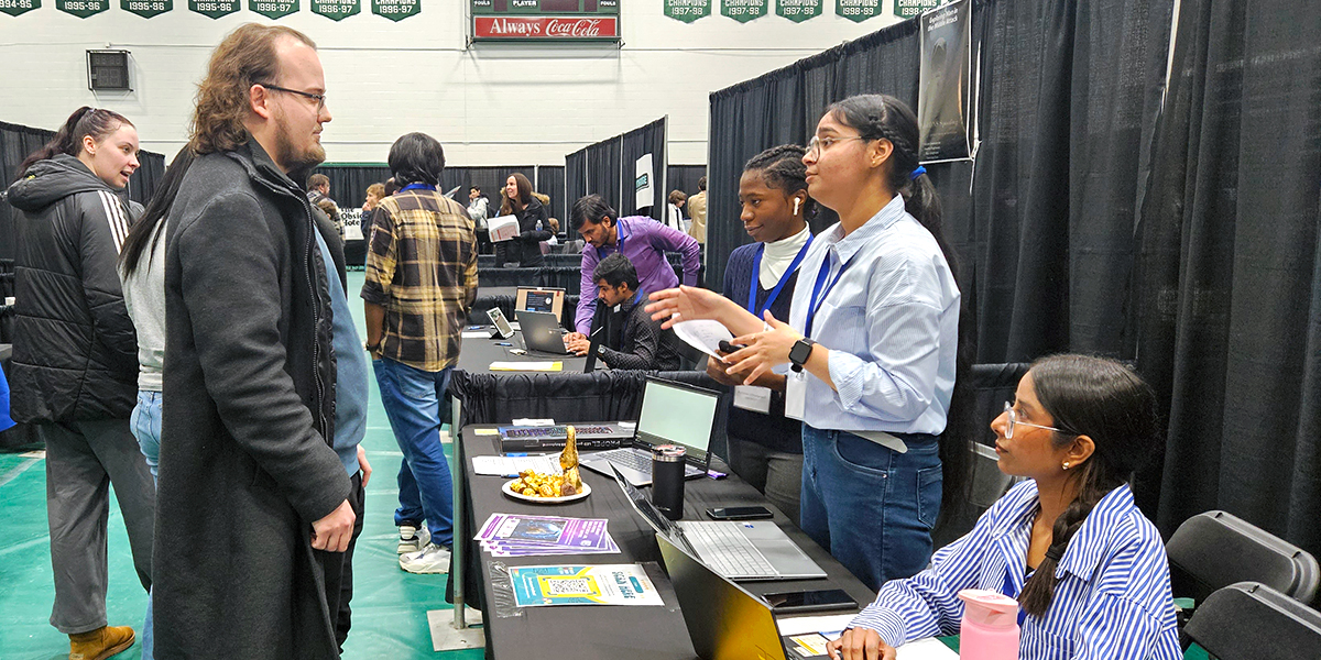 A man stands in front of a booth at the IT Student Expo. In the booth, a woman is gesturing and explaining a project.