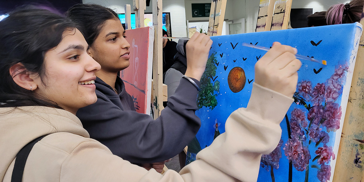 Two smiling students stand side-by-side and paint on a canvas.