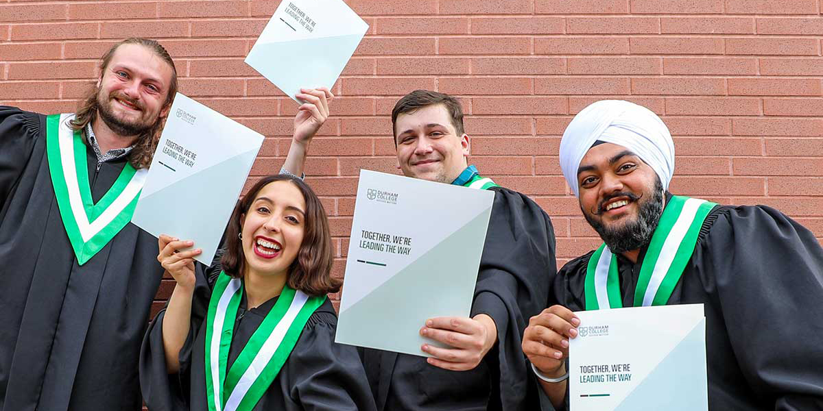 Four DC graduates in gowns hold up their diplomas and smile.
