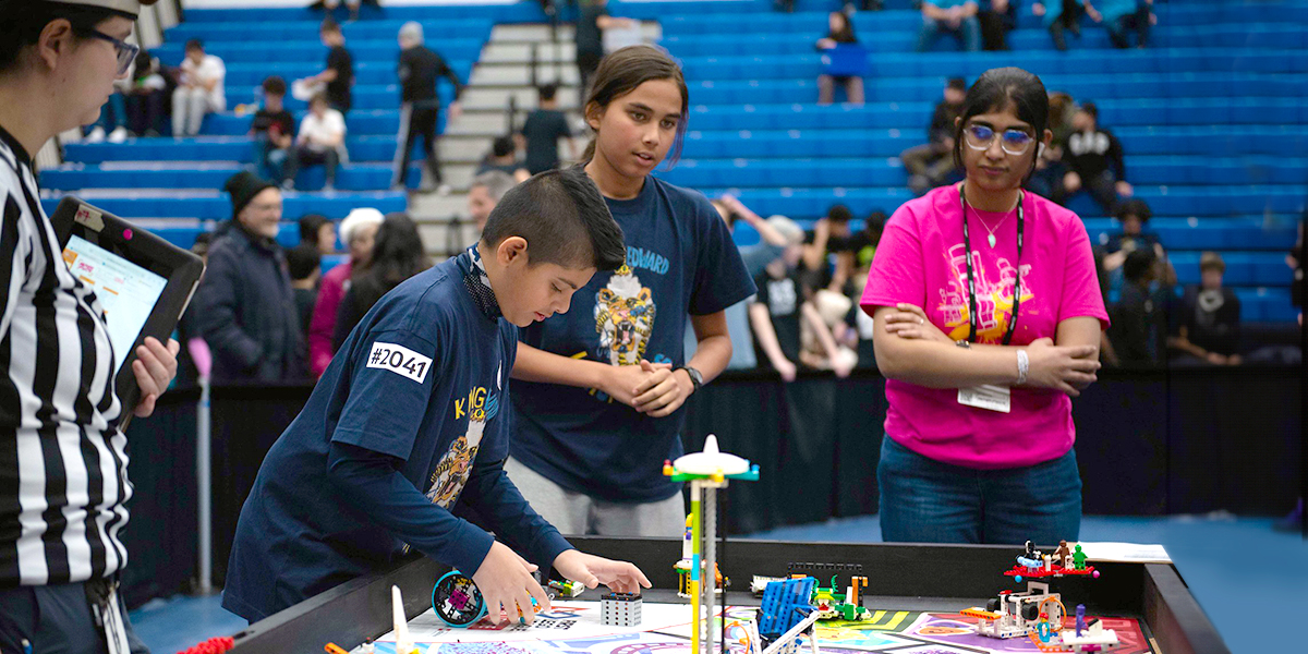 A competitor works on his Lego project while two other participants and a referee look on.