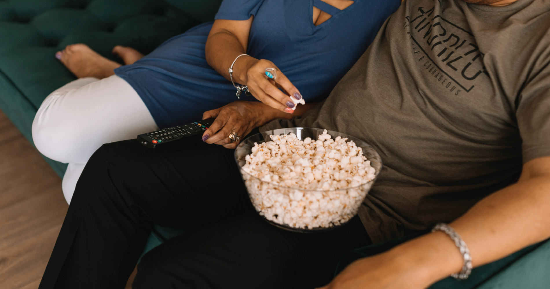 a couple sits comfortably on the couch, with a bowl of popcorn on a lap and the other person holds the remote