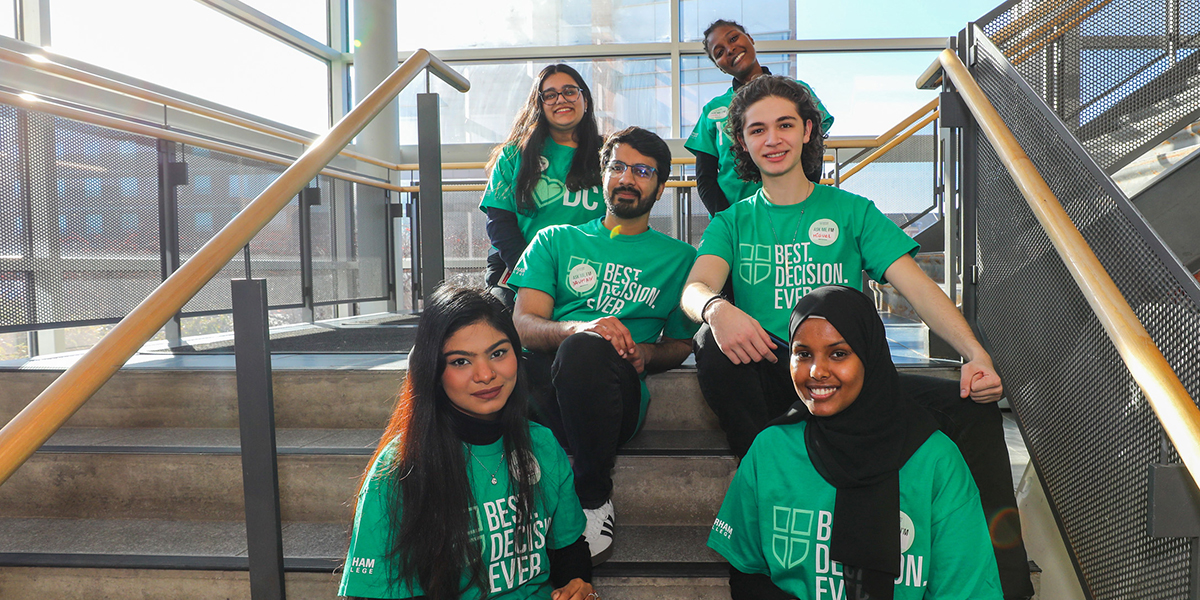 A group of students wearing Durham College green T-shirts sit on a staircase and smile at the camera.